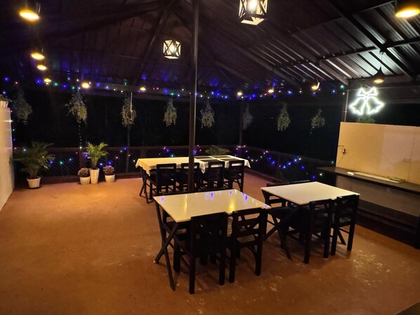 Un área de comedor al aire libre con mesas, sillas, luces colgantes y plantas en macetas.