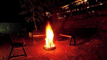 A campfire with two benches around it, set in a natural outdoor area with trees and a building in the background.