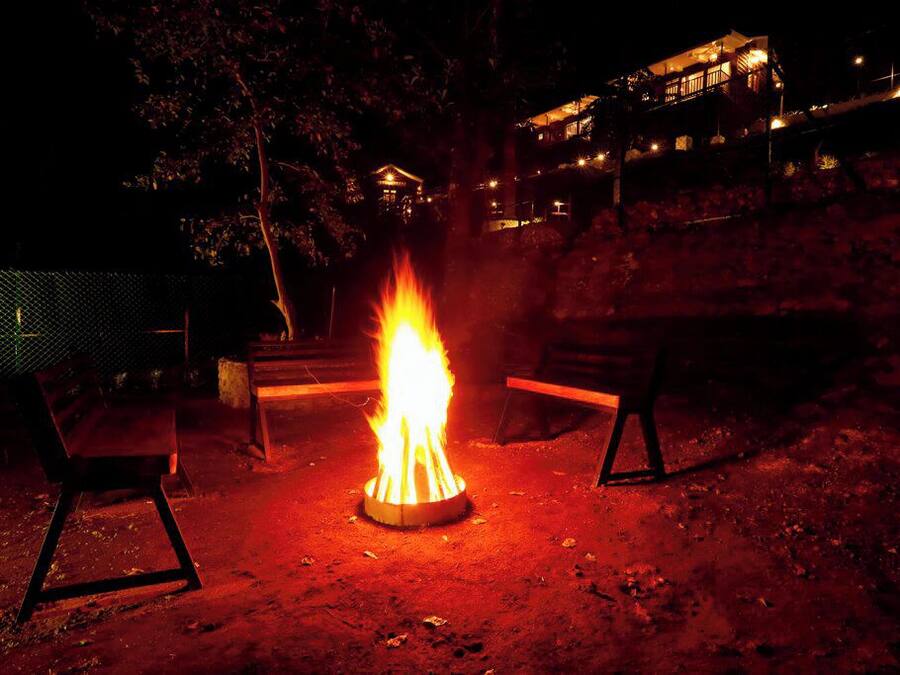 Ein Lagerfeuer mit zwei Bänken darum, eingebettet in eine natürliche Landschaft mit Bäumen und einem Gebäude im Hintergrund.