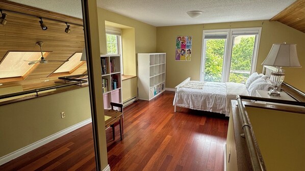 A bedroom with a large bed, a desk, a chair, a bookshelf, and a window with a view of greenery.