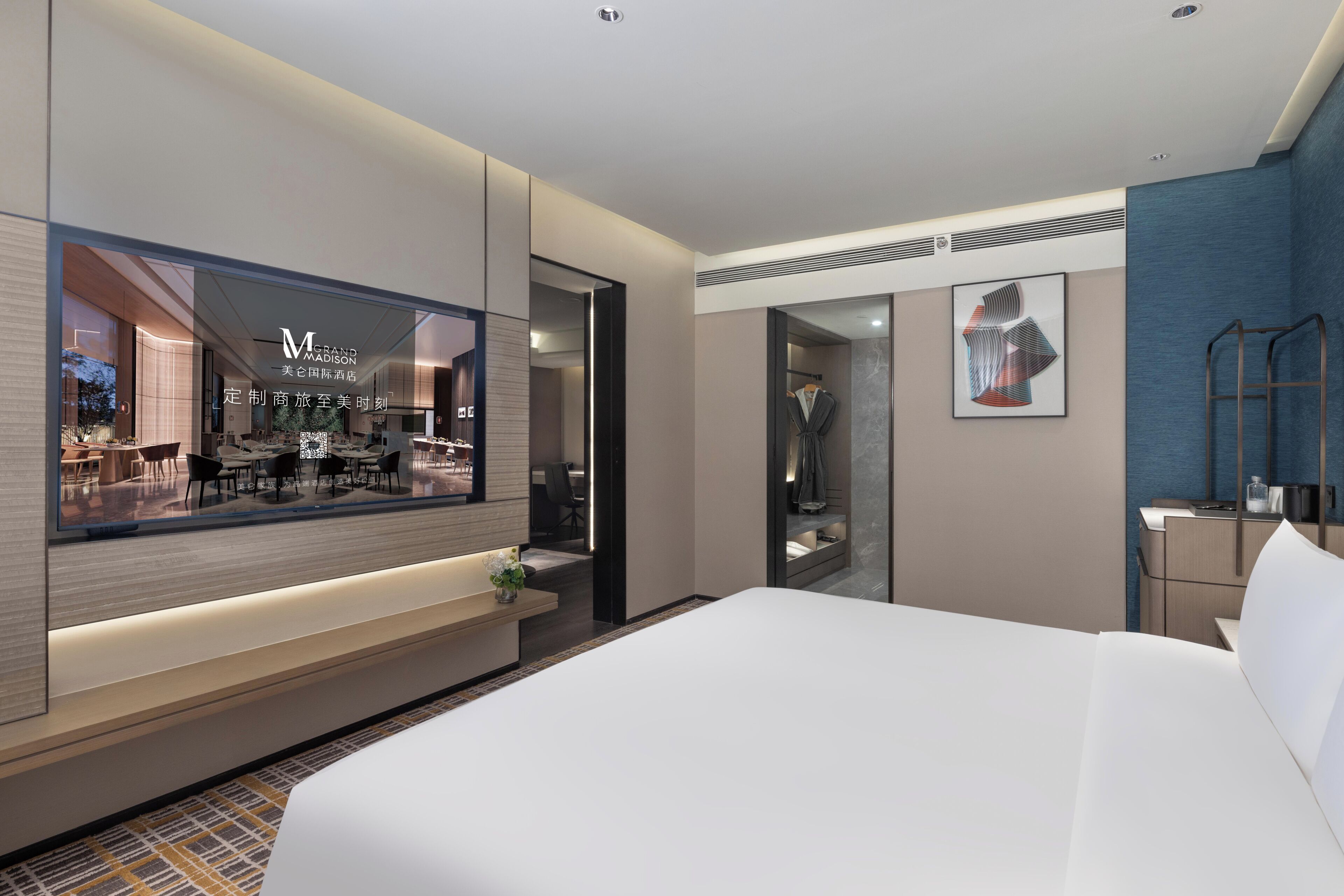 A modern hotel room with a large bed, a seating area with a bench, a desk, and a large window with a view of the hotel's interior.