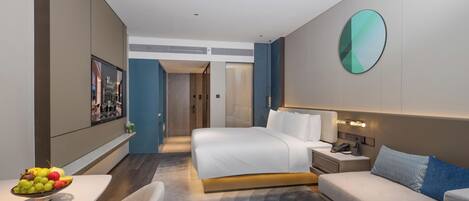 A modern hotel room with a bed, a sofa, a small table with fruit, and a wall-mounted TV.