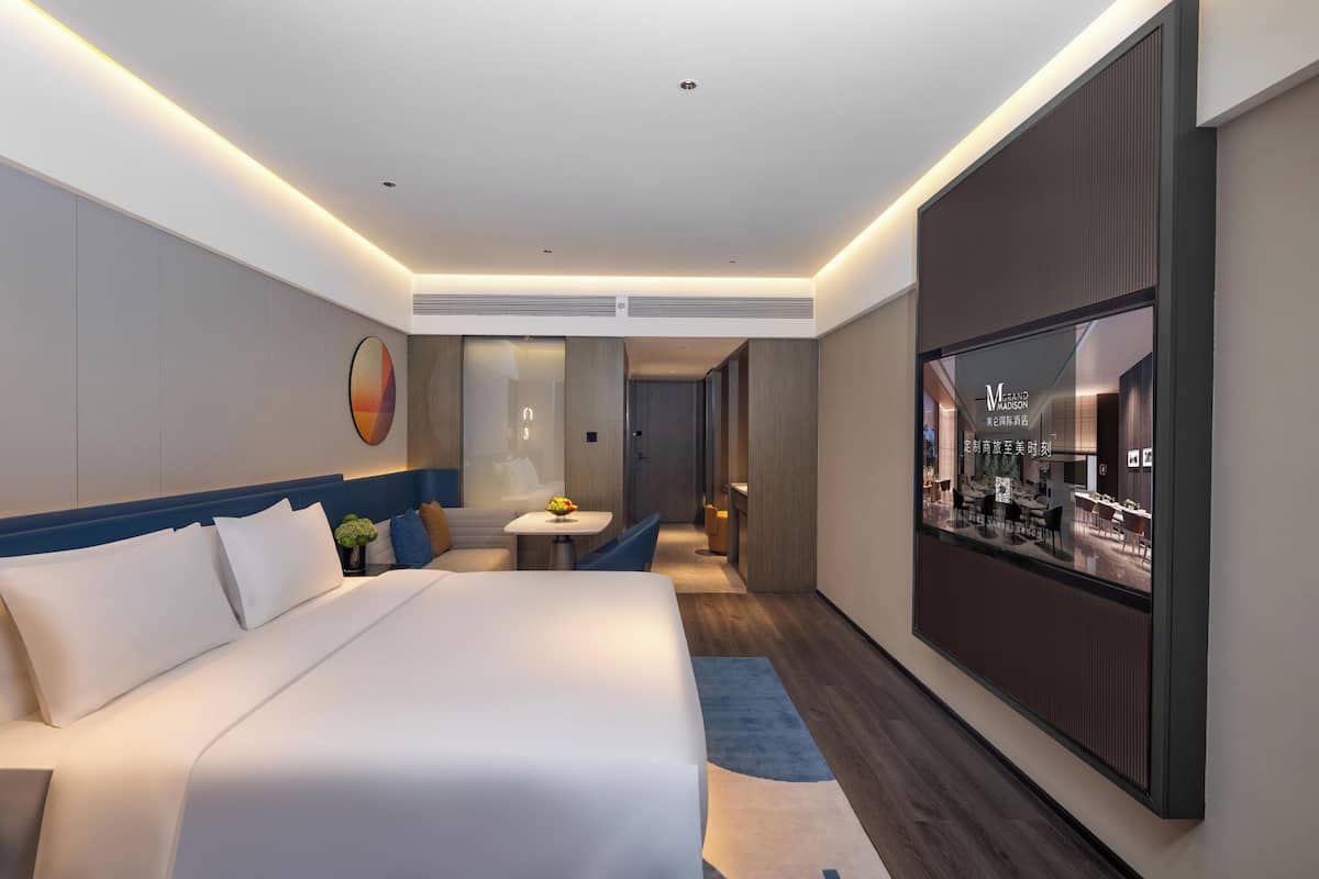 Une chambre d’hôtel moderne dotée d’un grand lit, d’une petite table avec un vase, d’un coin salon et d’une grande fenêtre donnant sur l’intérieur de l’hôtel.