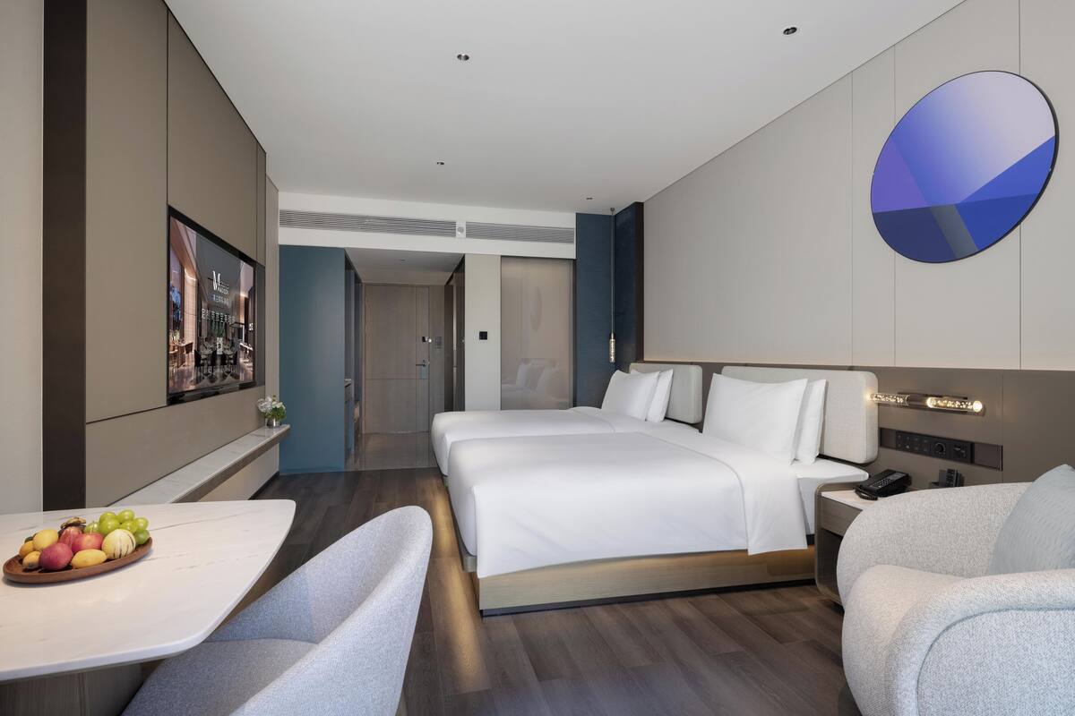 Une chambre d’hôtel moderne dotée d’un grand lit, d’un bureau avec un panier de fruits, d’un coin salon et d’un luminaire fixé au mur.