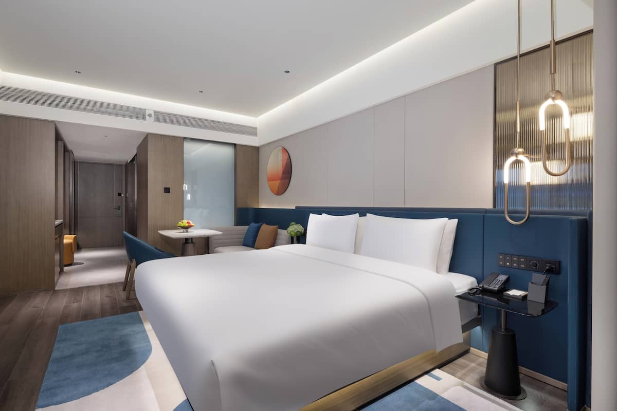 Une chambre d’hôtel moderne avec un grand lit, une table de chevet dotée d’un téléphone et d’un chargeur, et un luminaire fixé au mur.