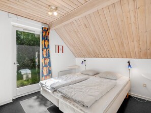 Une chambre avec un plafond en pente, un lit avec une literie blanche, une petite table de chevet et une fenêtre ornée d’un rideau coloré.