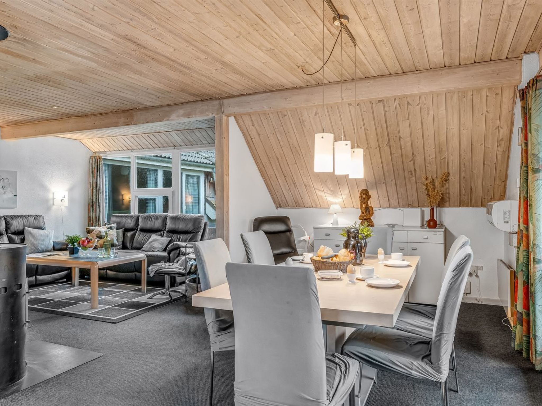 Een moderne, open woon- en eetruimte met een schuin plafond, houten balken en een groot raam dat voor natuurlijk licht zorgt.