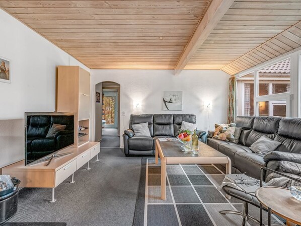 A modern living room with a wooden ceiling, a black leather sofa, a grey sofa, a wooden coffee table, and a TV on a wooden stand.