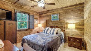 Een slaapkamer met een bed, houten wanden, een plafondventilator, een televisie, een raam en een nachtkastje met een lamp.