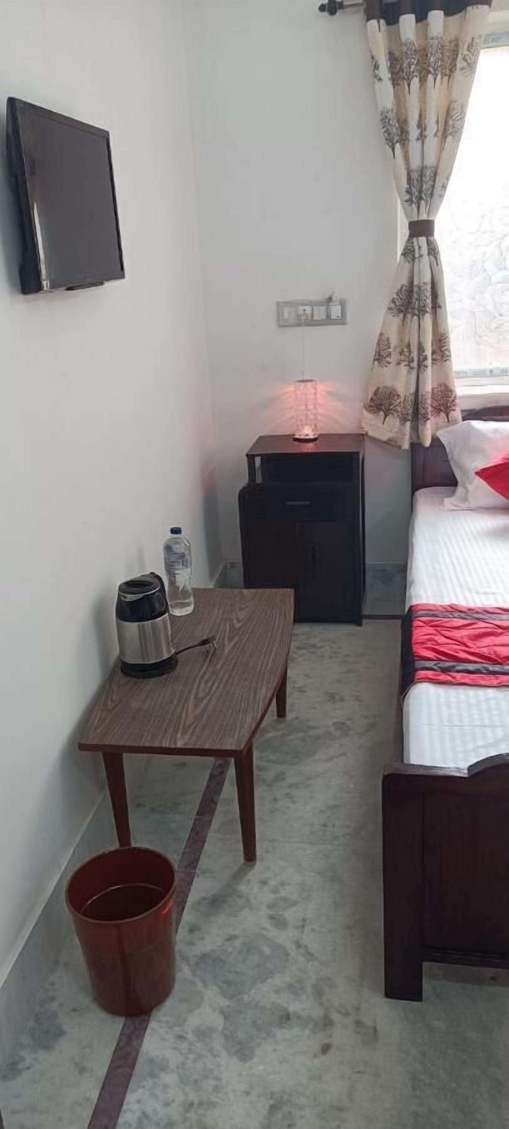 A small, neatly arranged room with a bed, a table with a kettle and a water bottle, a nightstand with a lamp, and a TV mounted on the wall.