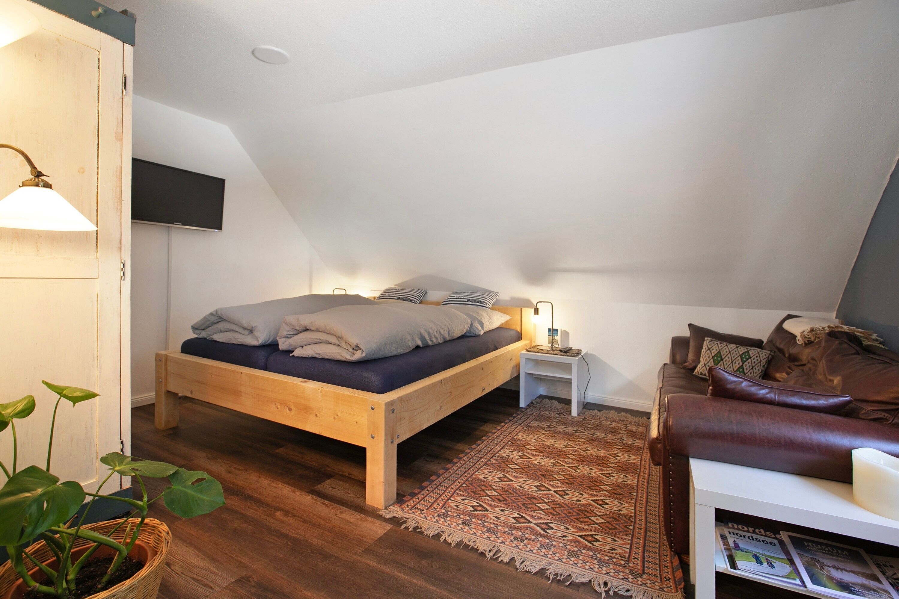 Een slaapkamer met een houten bed, een klein nachtkastje, een bruine leren bank, een kamerkleed met een patroon en een aan de muur bevestigde televisie.