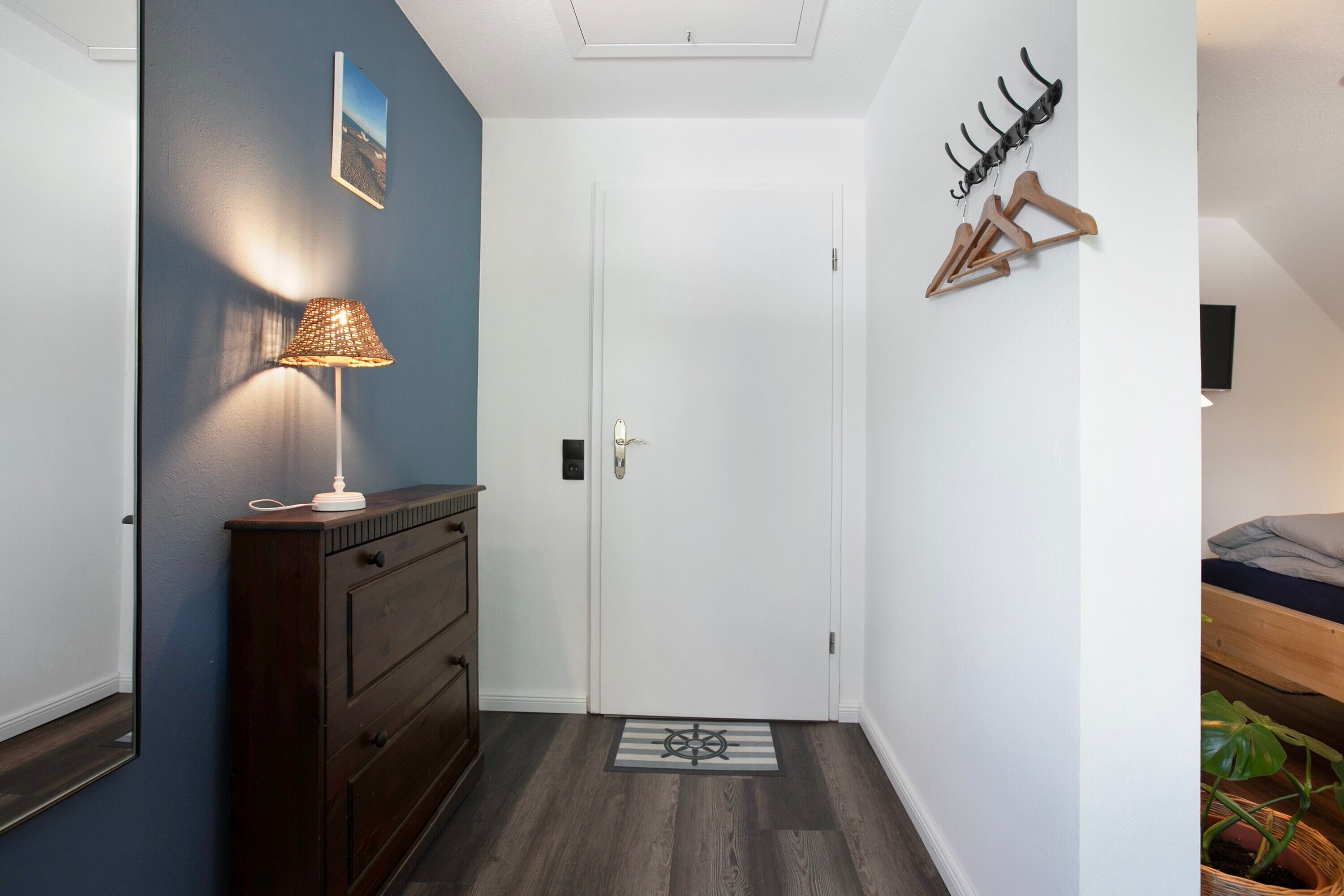 En hall med en byrå i trä, en lampa, en dörr med en dörrmatta och en klädhängare med galgar.
