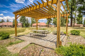 A wooden pergola with a picnic table in a landscaped outdoor area with houses in the background.