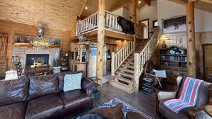 A rustic cabin interior with a wooden staircase, leather furniture, a fireplace, and a bookshelf.
