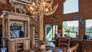 A rustic cabin interior with a dining area, wooden furniture, and antler chandeliers.