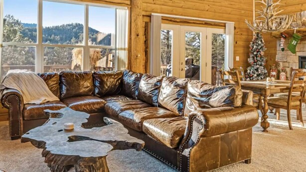 A wooden cabin interior with a leather sectional sofa, a rustic coffee table, and a Christmas tree.