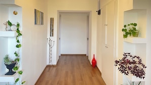 Un couloir avec un plancher en bois, des murs blancs et un lustre. On y trouve des étagères avec des plantes et une porte au fond.