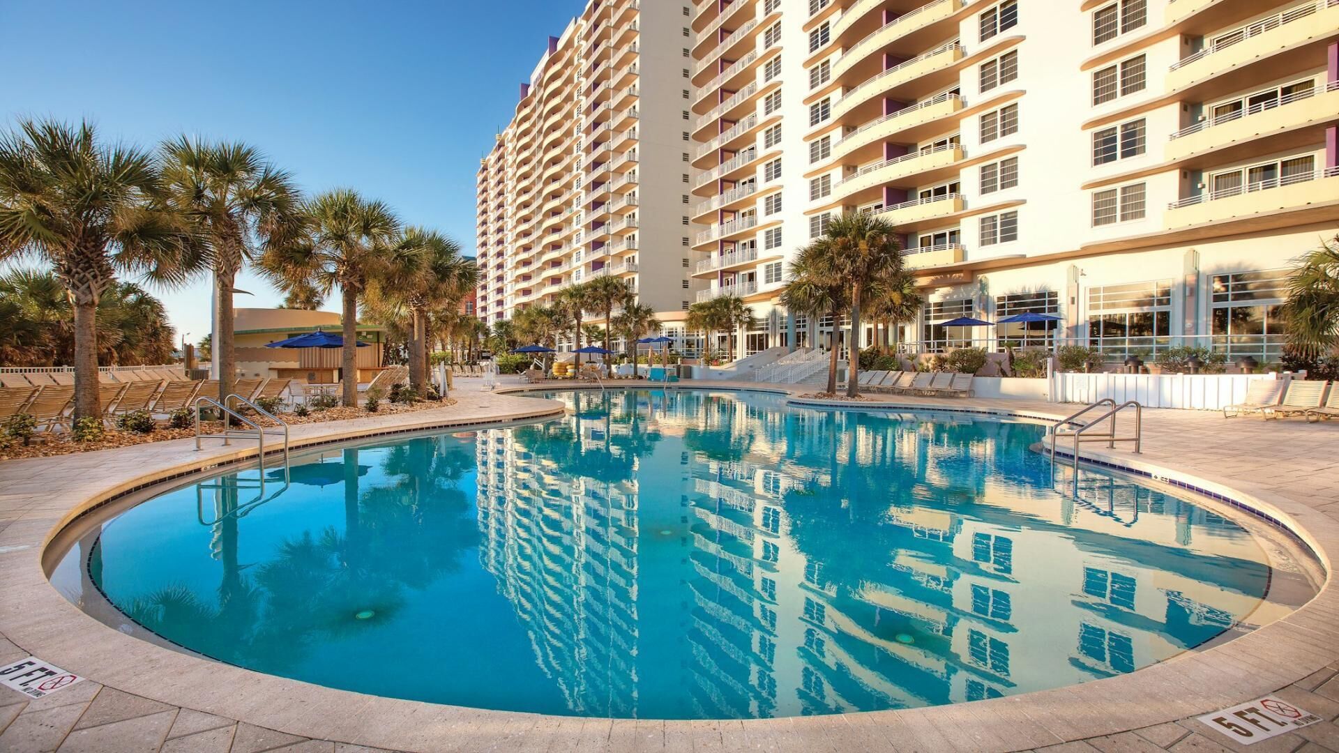 A large swimming pool surrounded by palm trees and a multi-story hotel building with balconies.