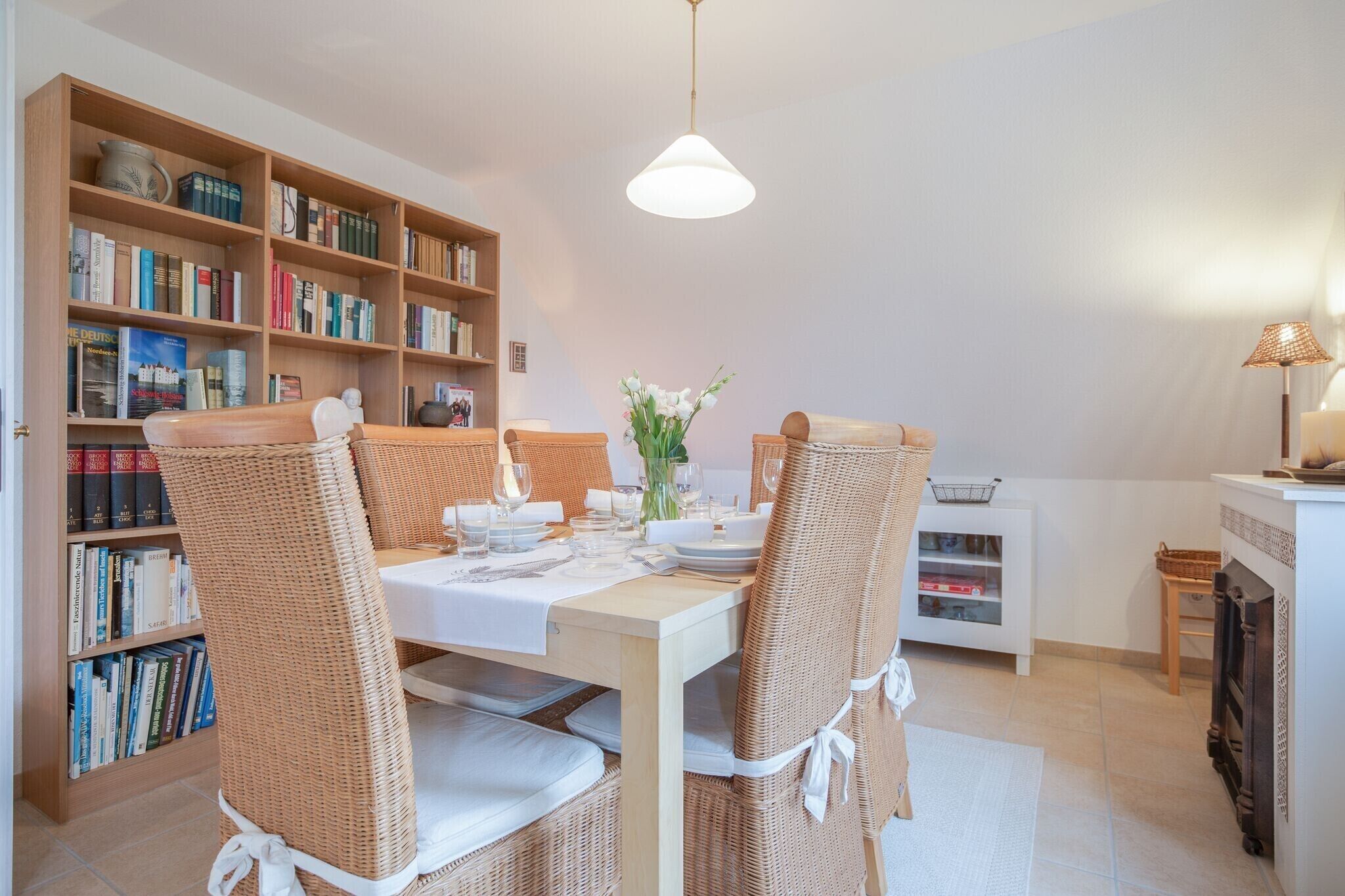 A dining area with a table set for four, surrounded by wicker chairs, a bookshelf, and a small side table with a lamp.