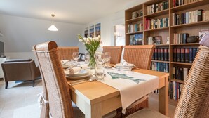 A dining area with a wooden table set for four, surrounded by wicker chairs, a vase with flowers, and a bookshelf filled with books. - Apartment 'Ferienwohnung Lillebror' with Shared Terrace, Shared Garden and Wi-Fi (Olderup)