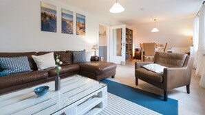 A living room with a brown leather sofa, a white coffee table, and a blue rug. There are two paintings on the wall, a dining area with a table and chairs, and a bookshelf. - Apartment 'Ferienwohnung Lillebror' with Shared Terrace, Shared Garden and Wi-Fi (Olderup)