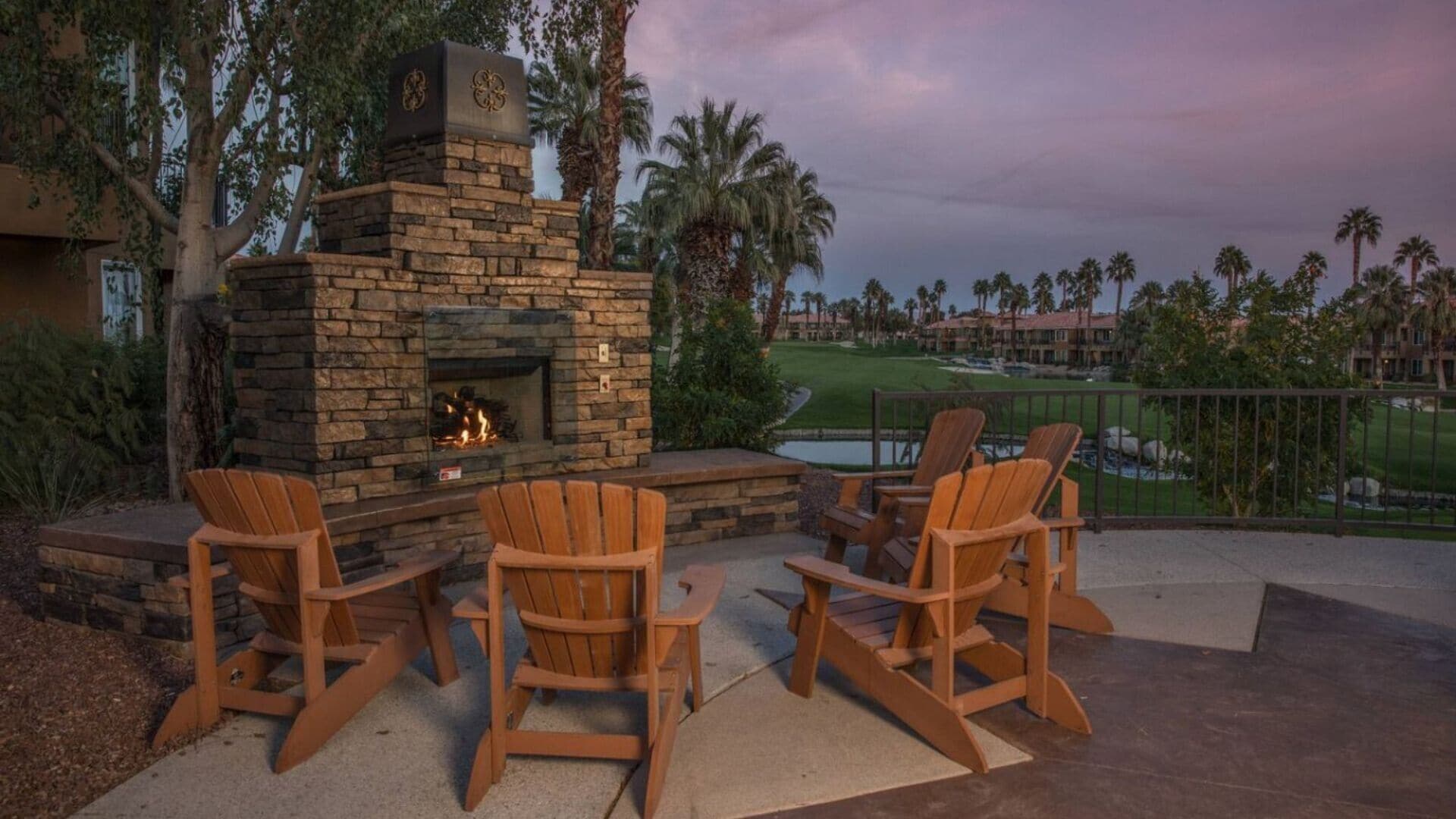 Une cheminée en pierre avec un feu, entourée de fauteuils Adirondack en bois sur une terrasse offrant une vue sur un terrain de golf et un bâtiment en arrière-plan.