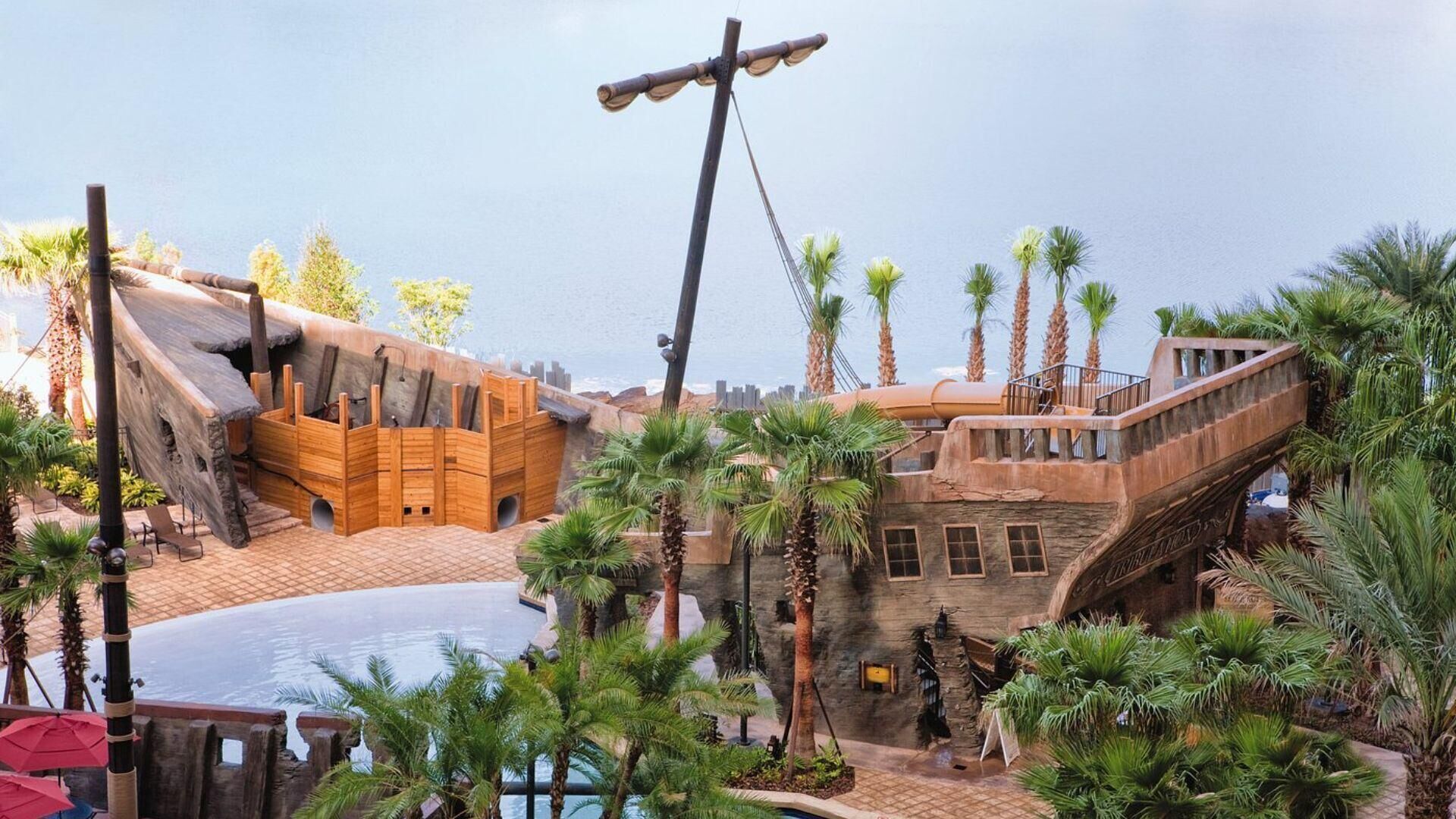 A tropical resort with a large pool, wooden structures resembling a ship, and palm trees.