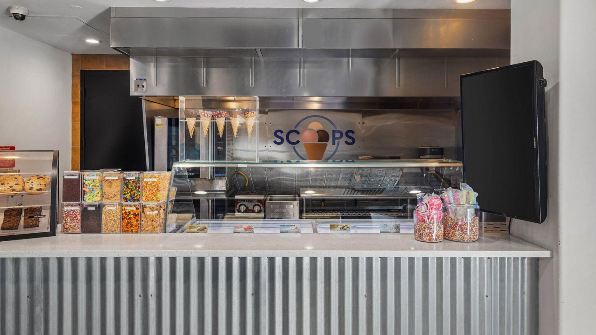 A dessert counter with various sweets and ice cream cones, a stainless steel hood above, and a 'SCOPS' logo.