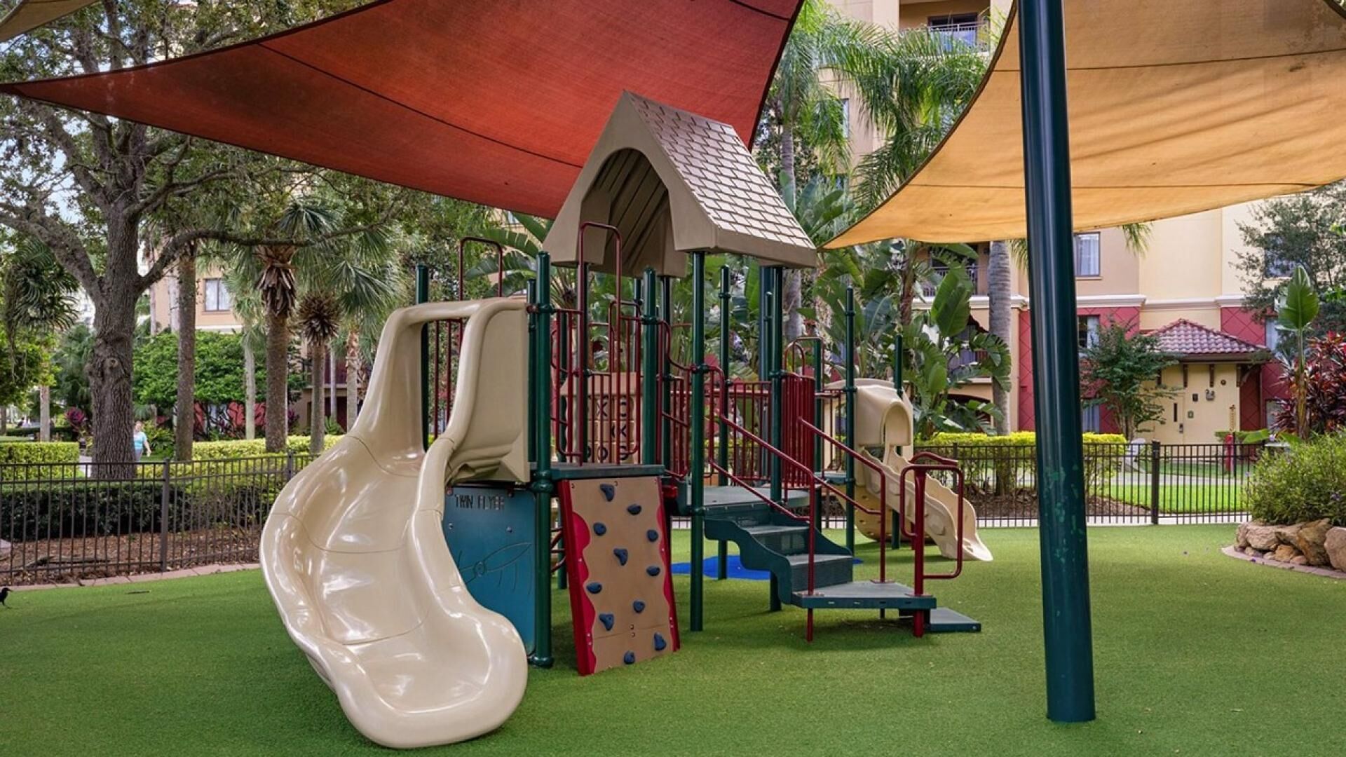 A playground with slides and climbing structures, surrounded by a fence and greenery.