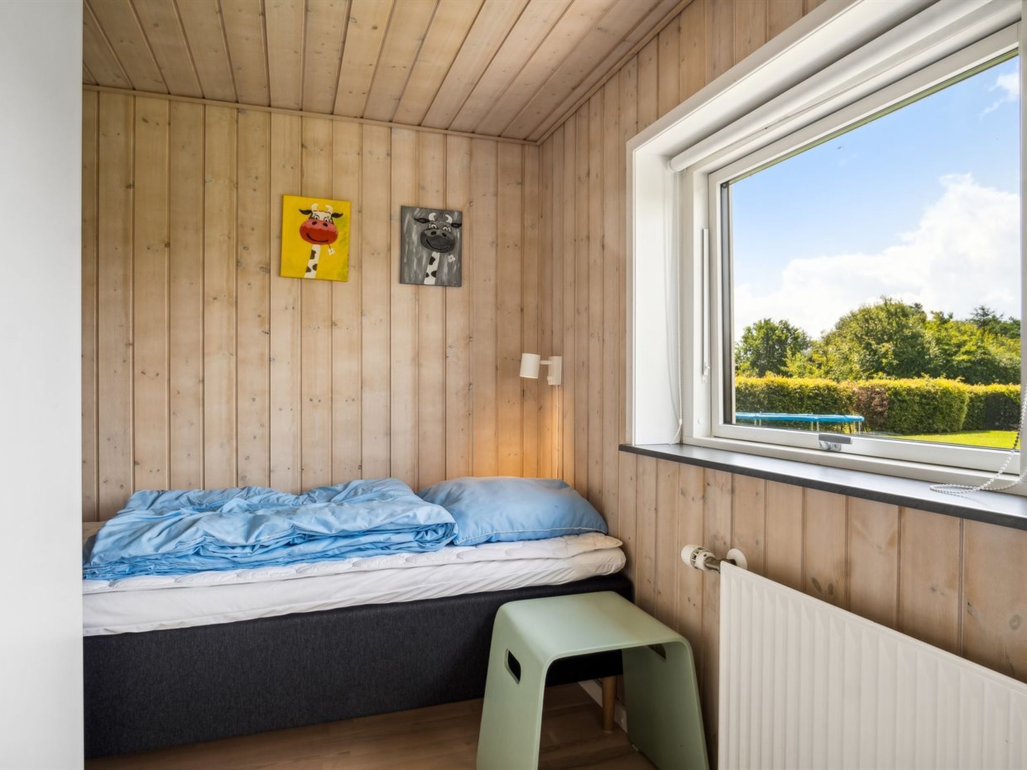 A bedroom with a bed, a small stool, a window with a view of greenery, and two wall-mounted artworks.