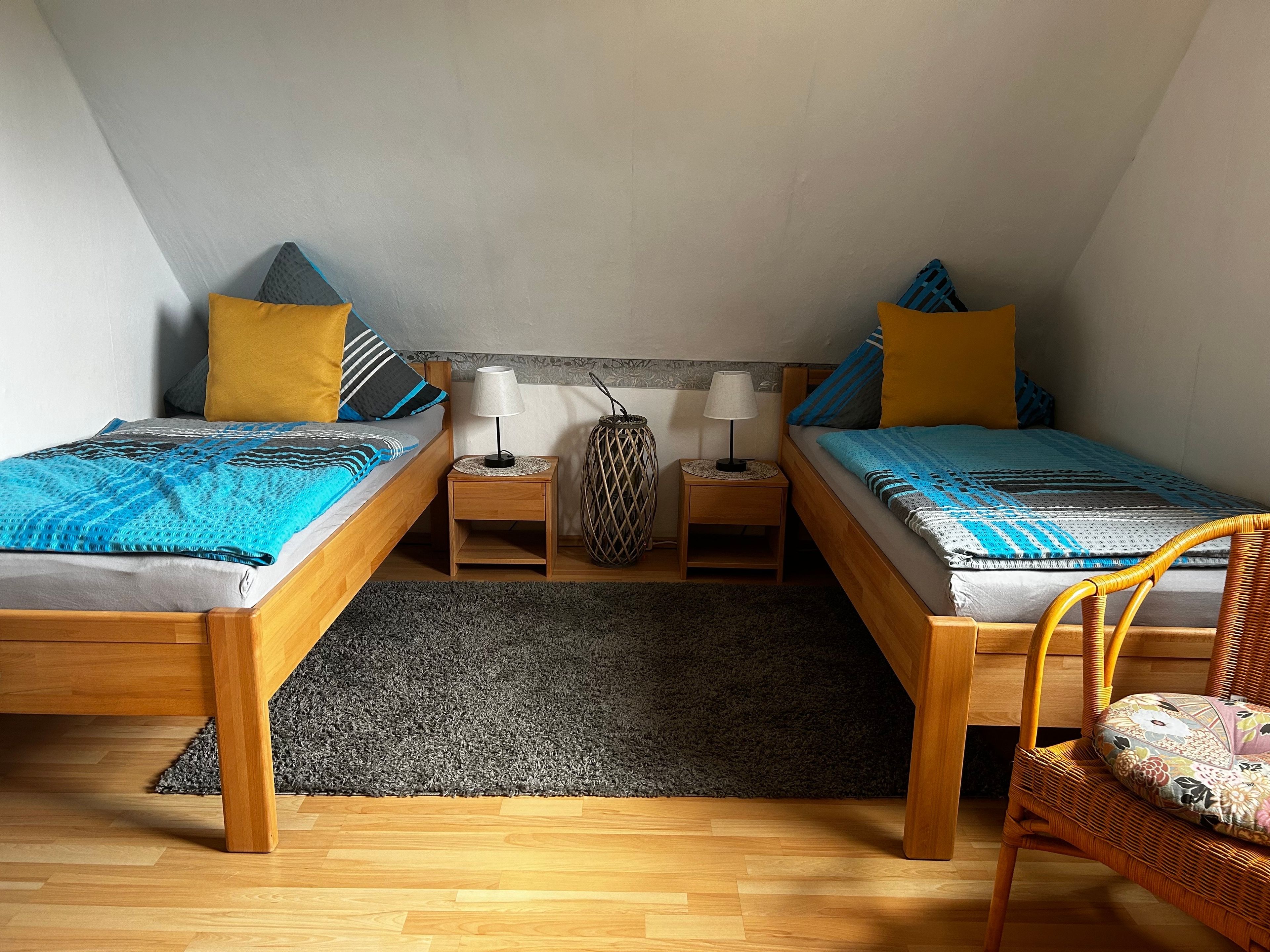 Ein kleines, gemütliches Schlafzimmer mit zwei Holzbetten, einem grauen Teppich und einem Korbstuhl.