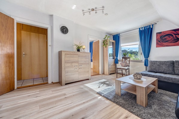 A modern living room with a wooden floor, a sofa, a coffee table, and a view through a window with blue curtains.