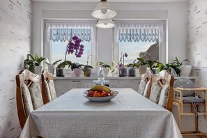 A dining area with a table, chairs, a fruit bowl, potted plants, and a window with curtains.
