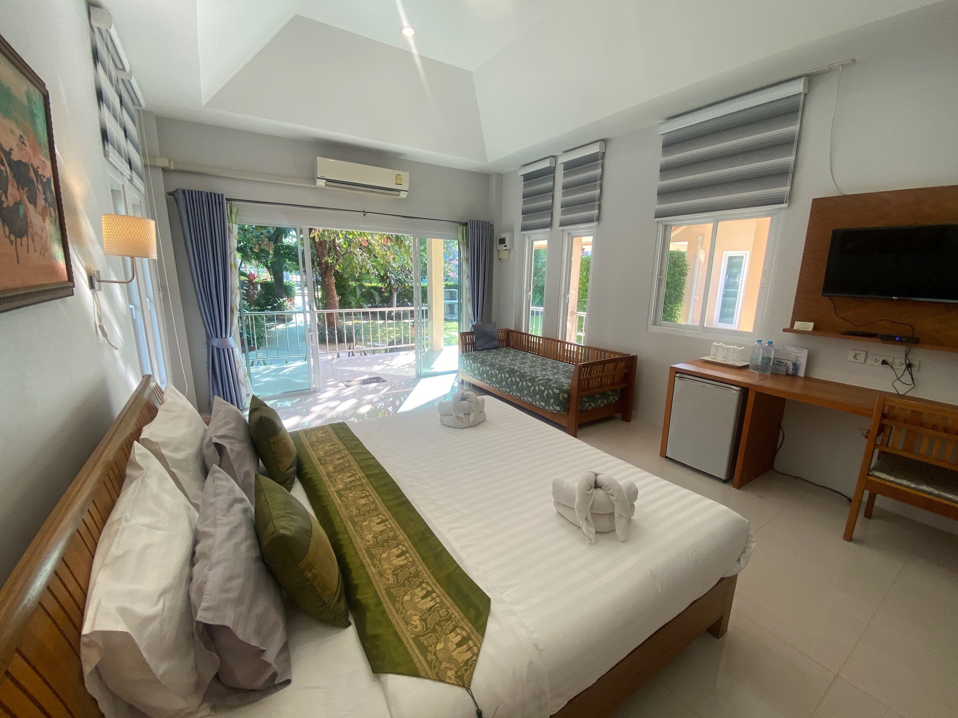 A hotel room with a large bed, a desk, a TV, and a balcony with a view of greenery.