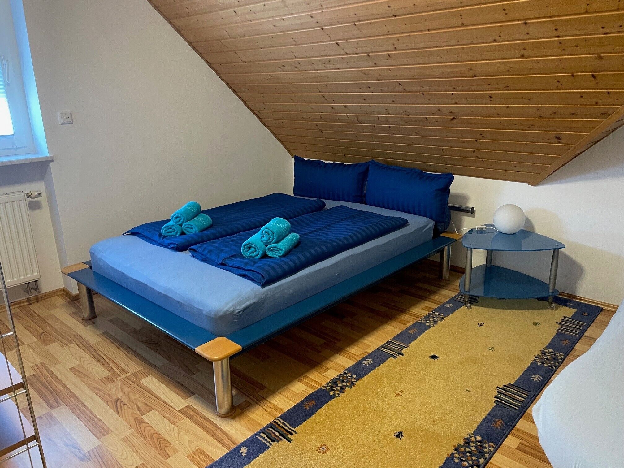 A single bed with blue bedding and pillows, a small bedside table with a lamp, a wooden floor, and a sloped ceiling.
