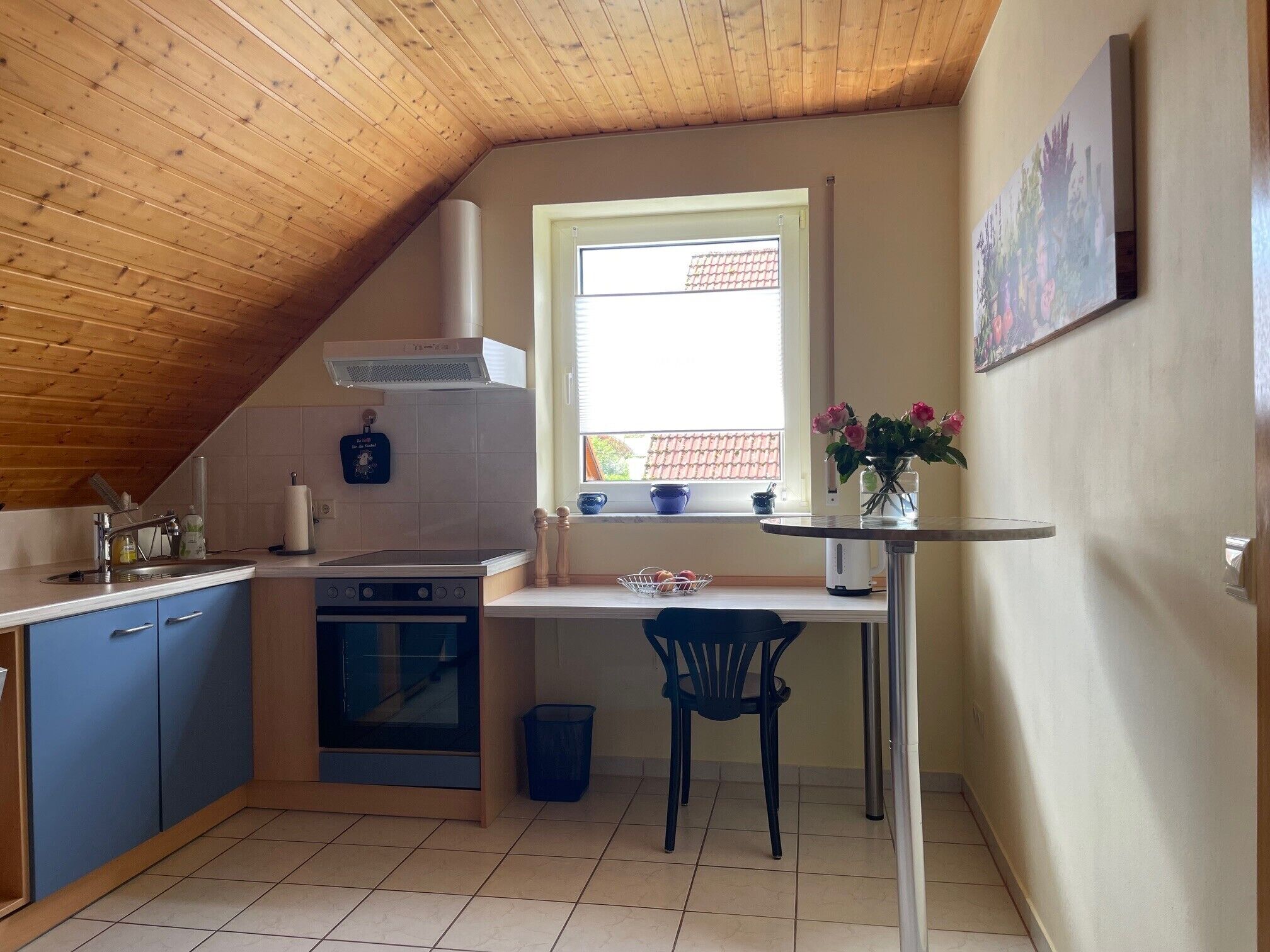 A kitchen with a stove, sink, and cabinets, a window with a view, and a painting on the wall.