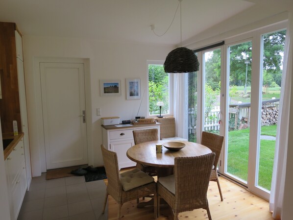 A dining area with a round table, wicker chairs, a ceiling light, and a view of a garden through large windows.