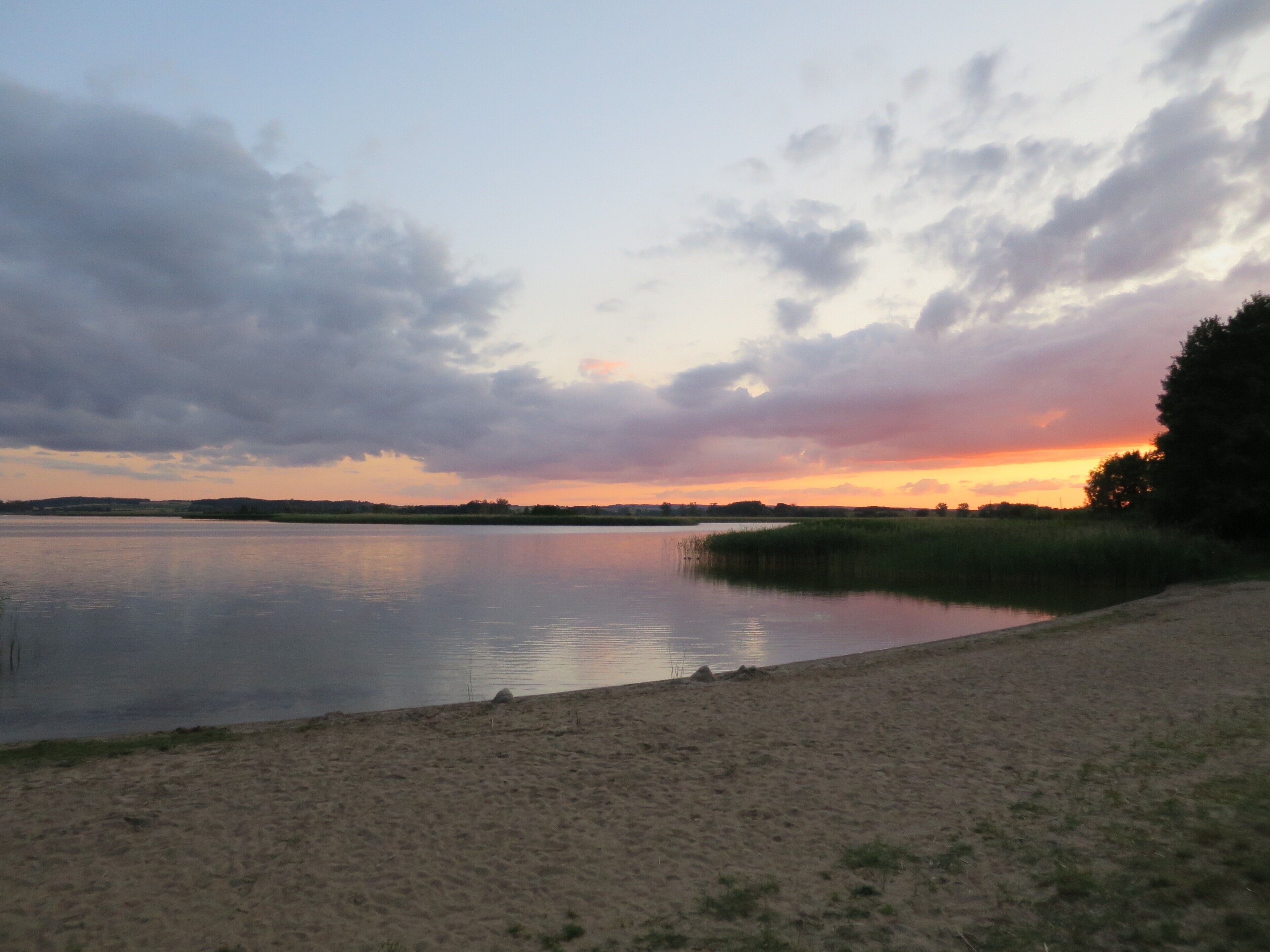 A calm lake with a sandy shore and a sunset in the background.