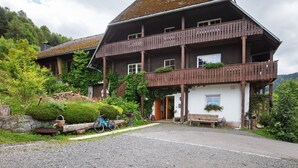 Una casa in legno a due piani con un balcone, immersa nel verde e con un vialetto sterrato.