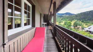 Un balcón de madera con un banco rojo, ventanas amplias y vistas a un valle boscoso.