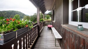 Una terraza de madera con un banco, una mesa, una ventana y vistas a una colina boscosa.