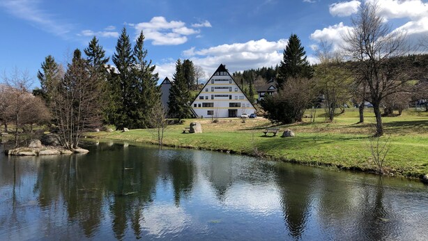 A multi-story building with a triangular roof, surrounded by trees and a body of water. - 2-room holiday apartment with sun terrace (Schönwald)