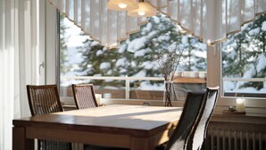 A dining area with a table, chairs, and a view of snow-covered trees through large windows. - 2-room holiday apartment with sun terrace (Schönwald)