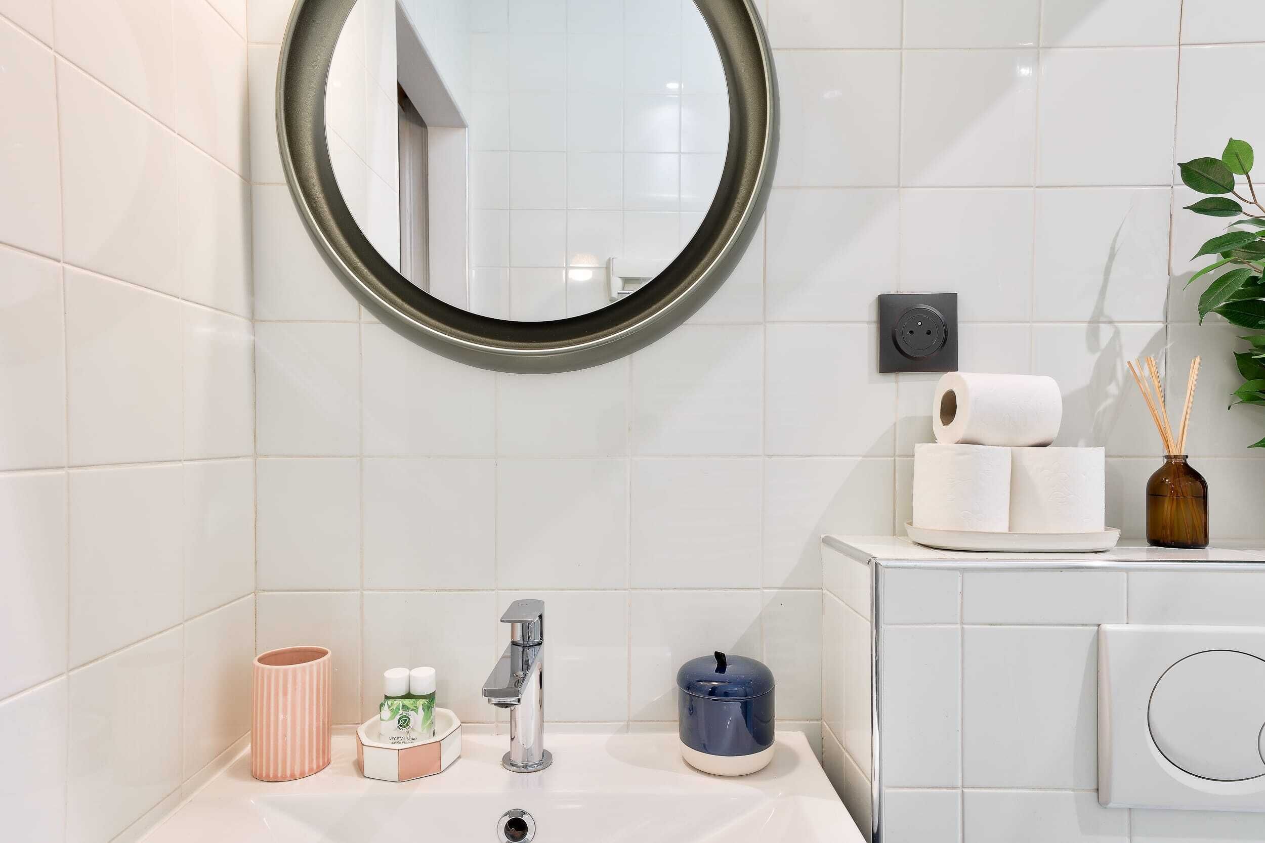 Une salle de bain avec un miroir rond, des carreaux blancs, un lavabo avec un robinet, un distributeur de savon, une bougie et une petite plante.