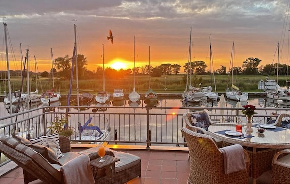 Une terrasse meublée de mobilier en osier, une table dressée pour le dîner et une vue sur les bateaux amarrés dans une marina au coucher du soleil.