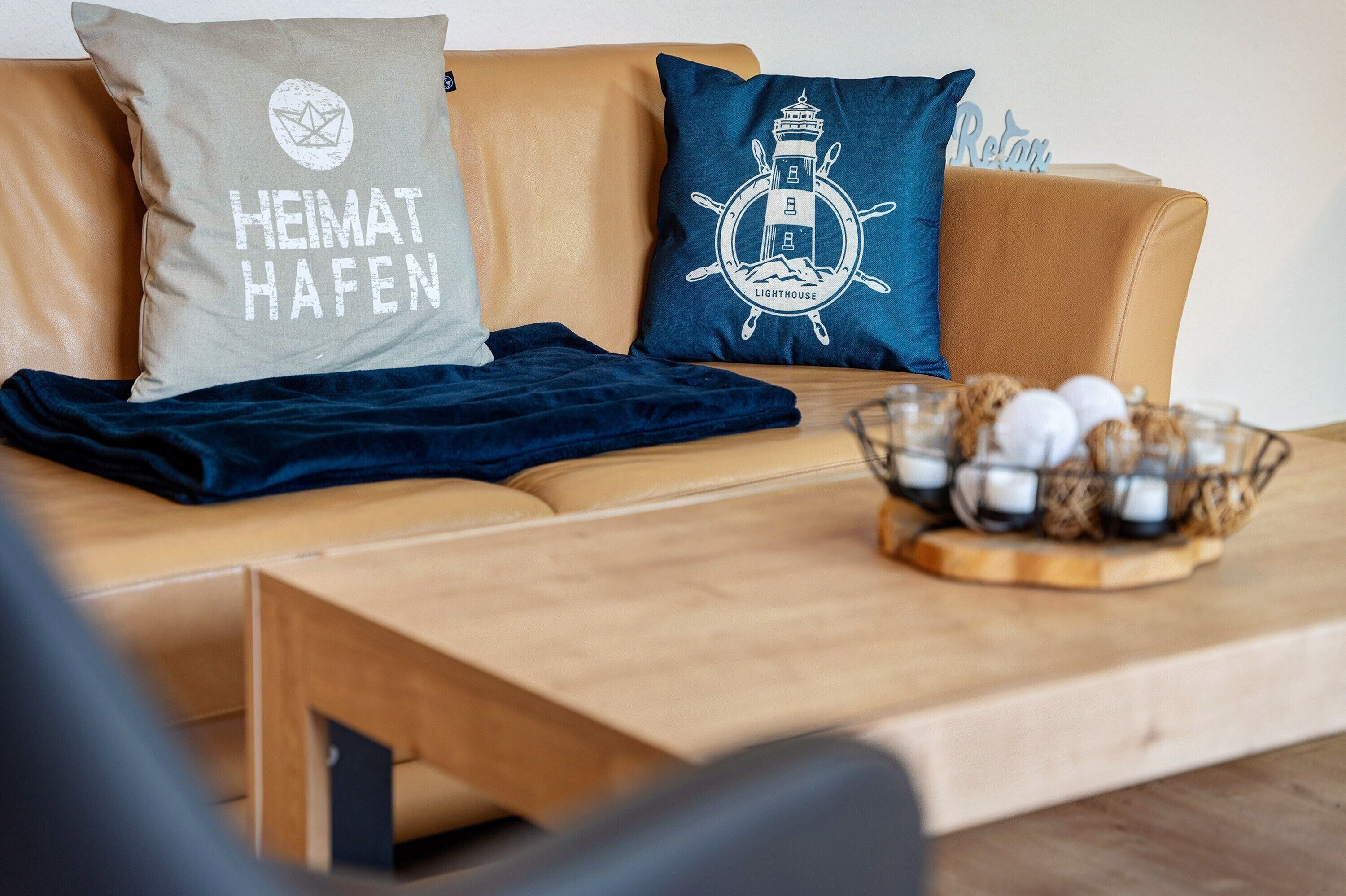 Een bank met twee kussens, waarvan één 'HEIMAT HAFEN' en de ander een ontwerp van een vuurtoren, en een houten bijzettafeltje met een mand vol flessen.