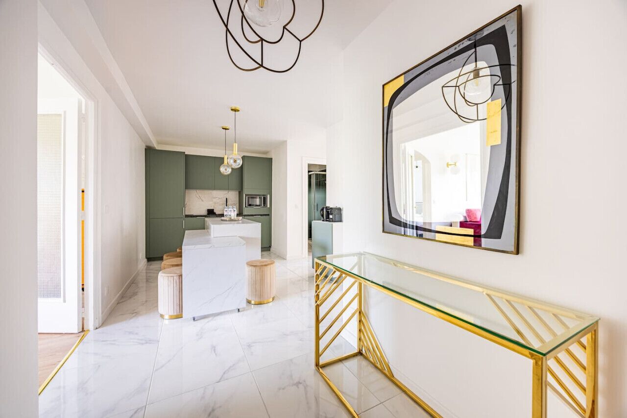 A modern interior with a glass console table, a large abstract mirror, and a kitchen in the background.
