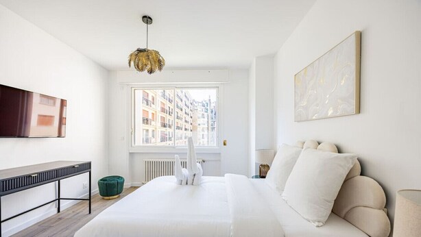 Une chambre moderne avec un grand lit, un bureau, un pouf vert, un radiateur, une fenêtre donnant sur des immeubles et un téléviseur fixé au mur.