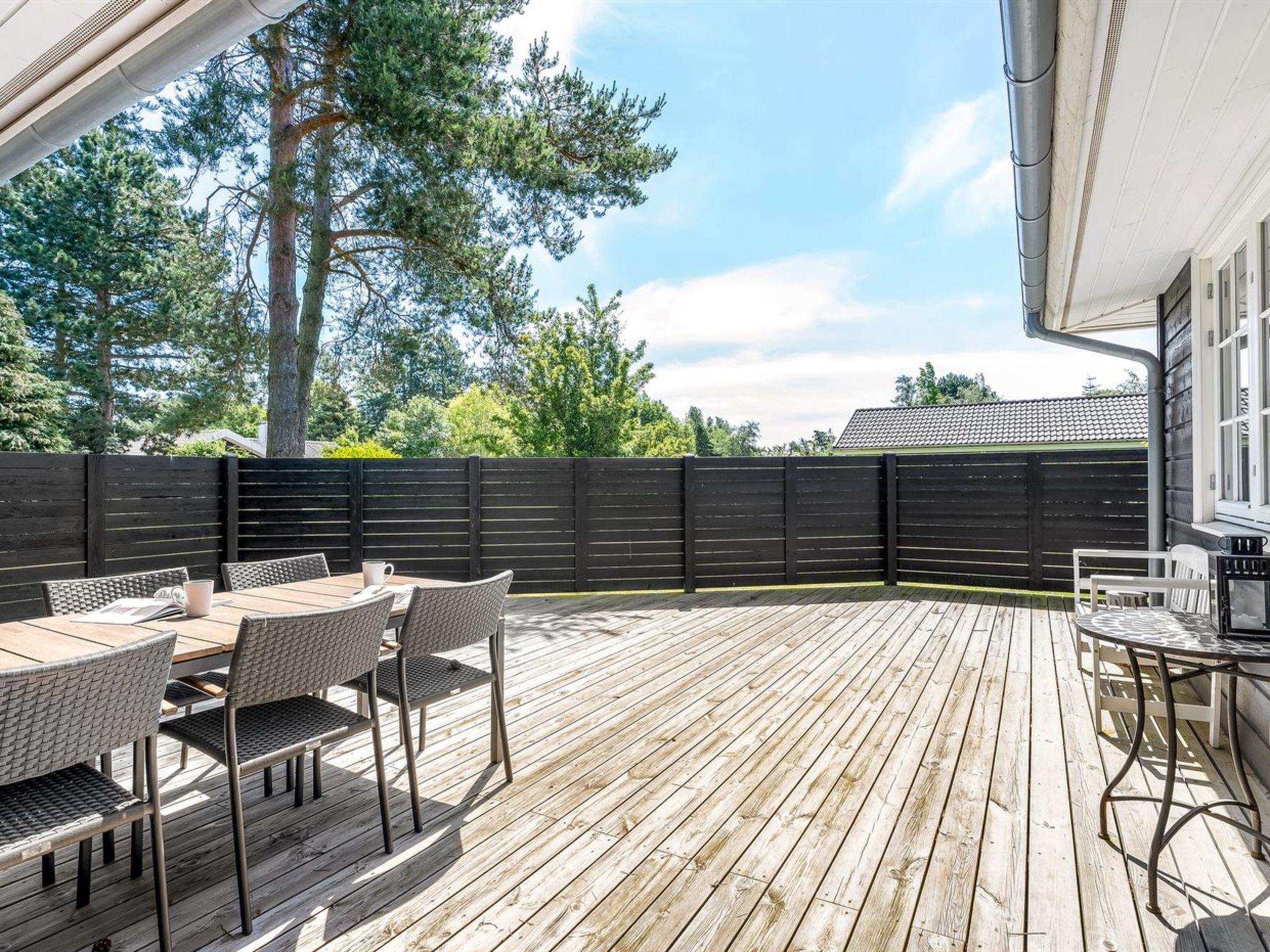 A wooden deck with outdoor furniture, a black fence, and a view of trees and a house in the background.