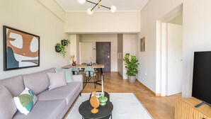 A modern living room with a grey sofa, a coffee table, a TV, and a dining area with chairs and a table. - Charismatic 2BR Apartment in Hilton (Athina)